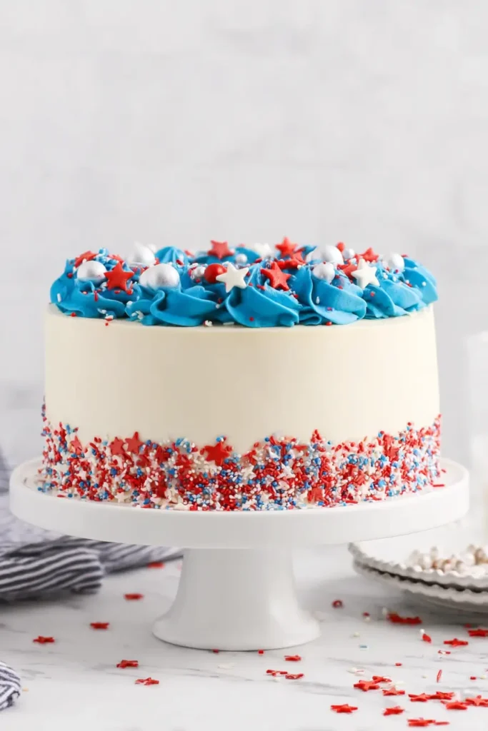 Patriotic 4th of July layer cake with smooth buttercream, piped red and blue swirls, and festive sprinkles on a white cake stand.