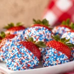 Close-up of strawberries dipped in white chocolate and red, white, and blue sprinkles on the bottom half, served on a white plate with checkered cloth.