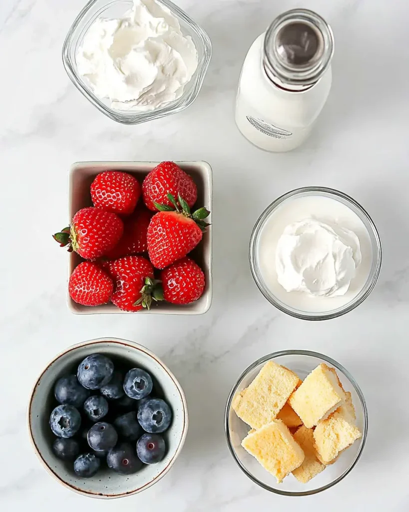 Overhead shot of 4th of July trifle ingredients including whipped topping, strawberries, blueberries, angel food cake cubes, milk, and pudding mix on white marble.
