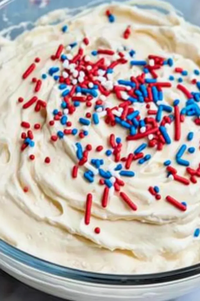 Red White and Blue Funfetti Dip – Easy No-Bake Party Treat 12 Red, white, and blue sprinkles sitting on top of creamy funfetti dip in a glass bowl before mixing.
