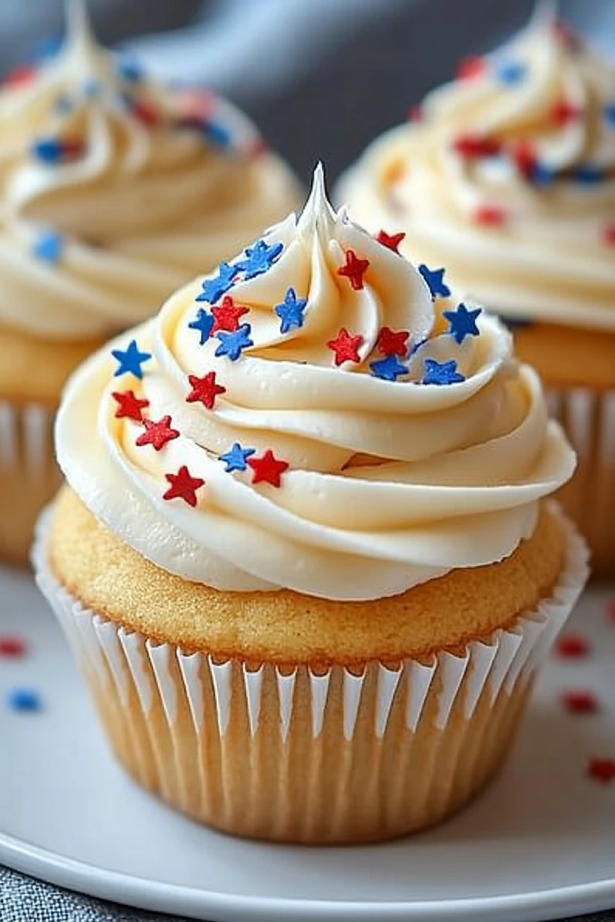 All-American Vanilla Cupcakes topped with creamy vanilla buttercream frosting and decorated with red and blue star-shaped sprinkles on a white plate with a soft gray linen background.