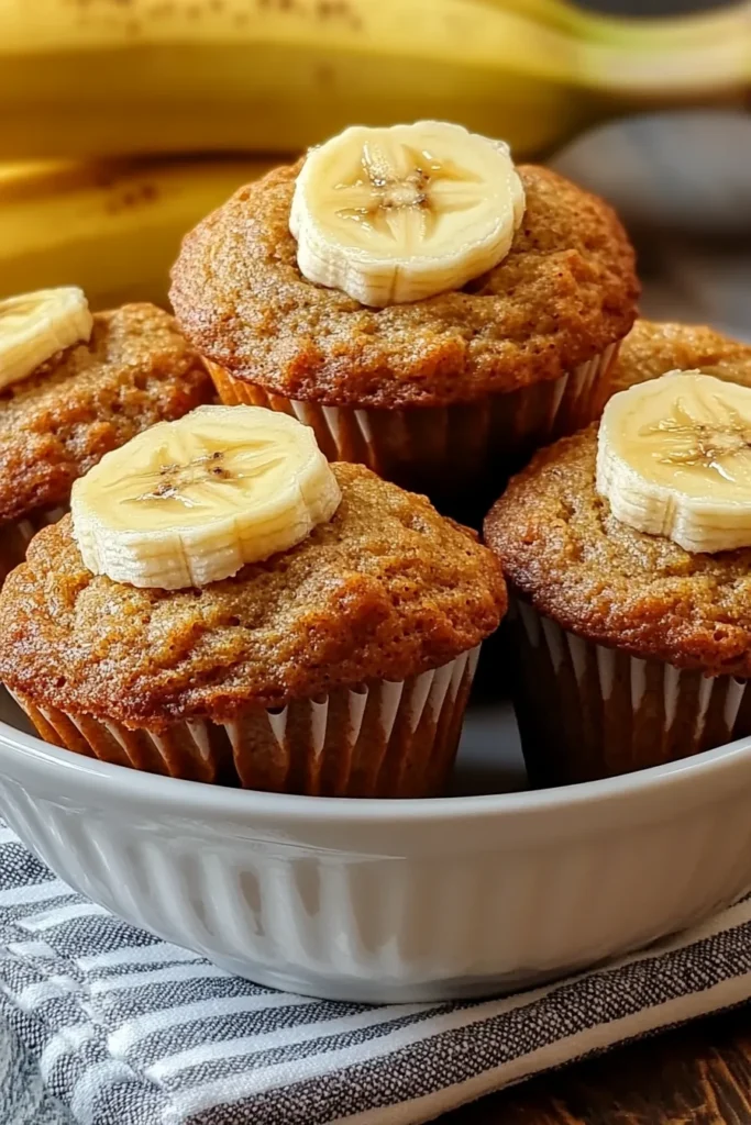 Banana Bread Muffins Recipe: Easy, Moist & Flavorful 8 Banana bread muffins recipe served in a white bowl, topped with banana slices and placed alongside ripe bananas on a rustic kitchen surface.