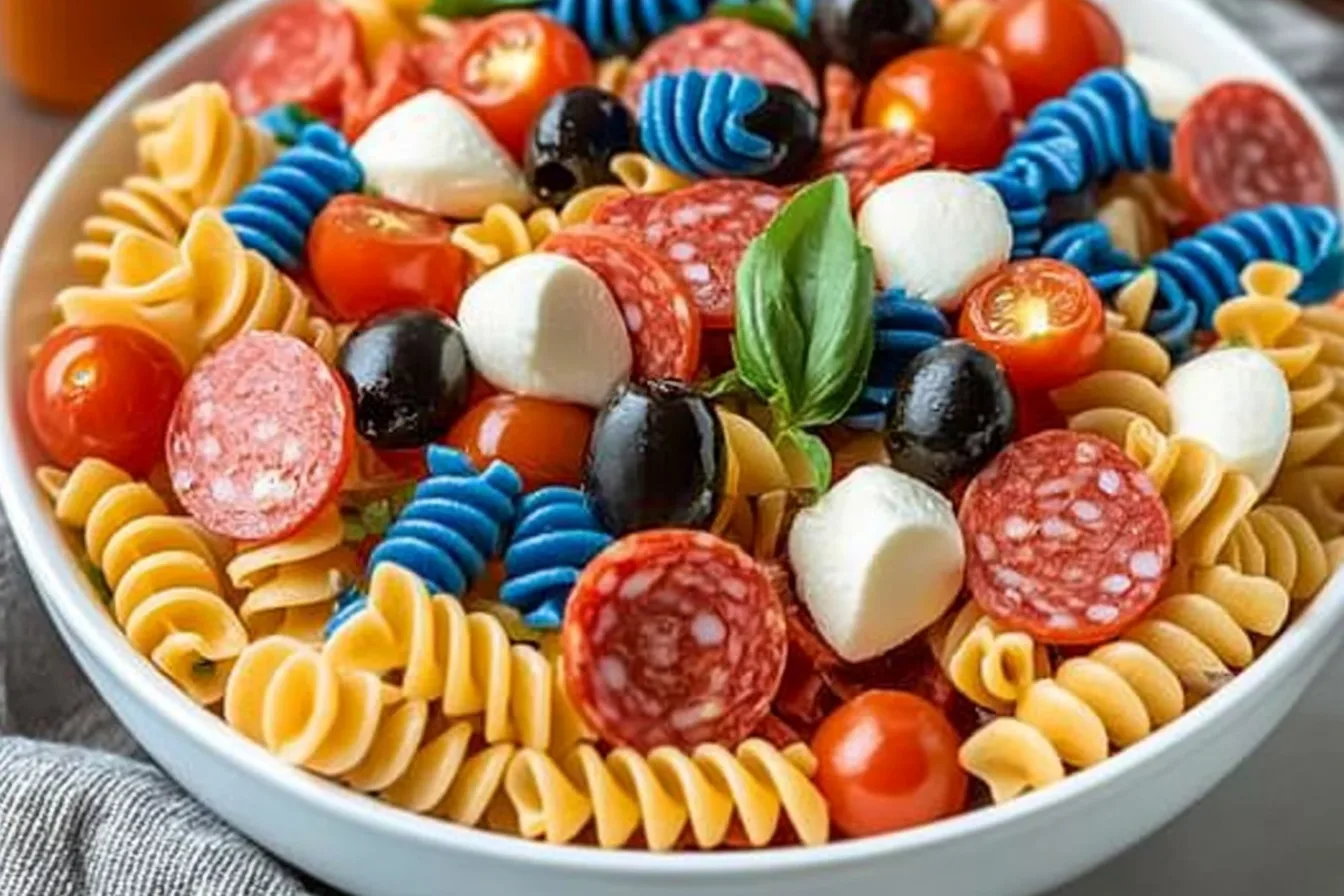 Chilled 4th of July pasta salad with red, white, and blue rotini, pepperoni, black olives, mozzarella, and cherry tomatoes in a white bowl.