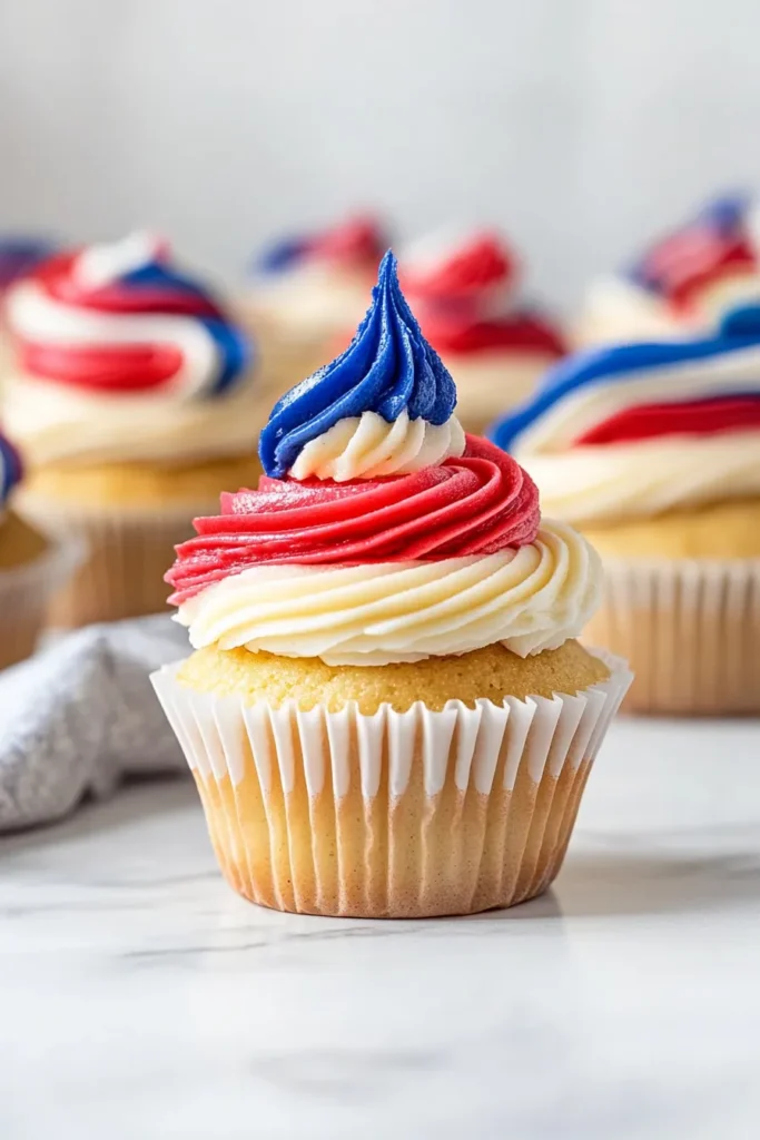 Close-up of a patriotic cupcake with red, white, and blue swirled buttercream frosting on a golden vanilla base, set on a white marble countertop with blurred cupcakes in the background.