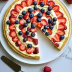 Top-down view of a patriotic 4th of July fruit pizza with a golden sugar cookie crust, creamy white cream cheese frosting, and fresh blueberries, strawberries, and raspberries arranged in concentric circles. A slice is served on a white ceramic plate on a rustic wooden table.