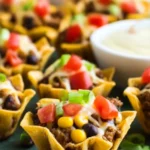 Baked Taco Bites Recipe: Easy & Flavorful Party Snack 6 Close-up of crispy baked taco bites in golden mini shells filled with seasoned ground beef, black beans, corn, melted cheese, and topped with fresh diced tomatoes, green onions, and cilantro on a rustic dark green slate.