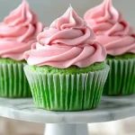 Delicious Fluffy Watermelon Cupcakes Recipe for Summer Fun 7 Three fluffy watermelon cupcakes with green cupcake bases and creamy pink frosting swirls displayed on a white marble cake stand against a soft beige background.