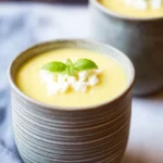 Delicious Fruity Summer Cold Pineapple Soup Recipe 7 Two handcrafted ceramic cups filled with creamy fruity summer cold pineapple soup, garnished with crumbled white cheese and fresh basil on a rustic teal wooden table with a dark navy fabric background.