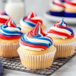 Patriotic cupcakes with red, white, and blue swirled buttercream frosting on vanilla bases, displayed on a wire cooling rack over a gray concrete table.