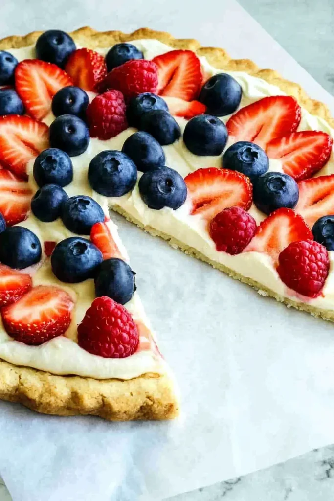 Close-up top-down view of a 4th of July fruit pizza with golden sugar cookie crust, creamy white frosting, and fresh blueberries, sliced strawberries, and raspberries, with one slice removed.