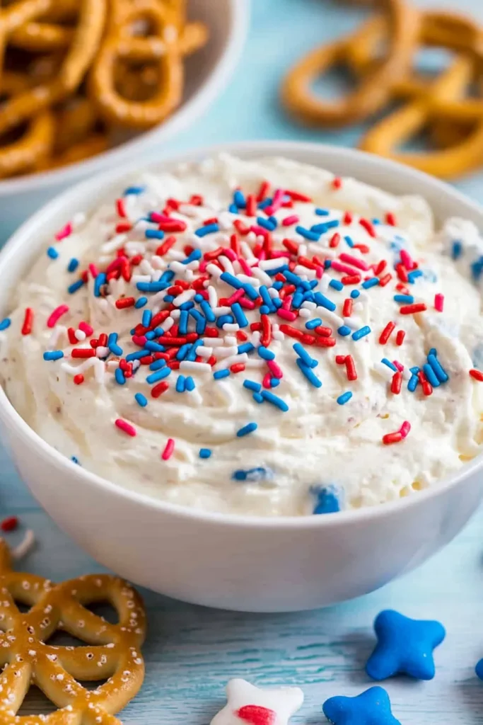 Red White and Blue Funfetti Dip – Easy No-Bake Party Treat 14 Finished Red, White and Blue Funfetti Dip in a white bowl topped with patriotic sprinkles, surrounded by vanilla wafers and pretzels.