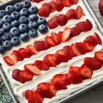 Flag Cake Recipe: Easy Strawberry Jello Dessert for July 4th 9 Patriotic flag cake decorated with strawberries and blueberries on whipped cream in a metal pan.