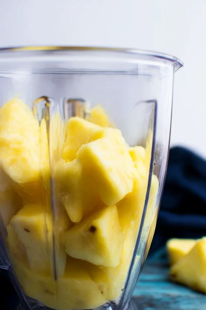 Delicious Fruity Summer Cold Pineapple Soup Recipe 6 Fresh yellow pineapple chunks in a clear blender container, ready to be blended for a tropical cold pineapple soup, with pineapple slices on a white kitchen countertop.