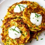 Easy Zucchini Fritters Recipe: Crispy & Healthy Summer Snack 8 Golden crispy zucchini fritters topped with creamy sour cream and fresh chopped chives on a white ceramic plate, set against a light gray textured surface.