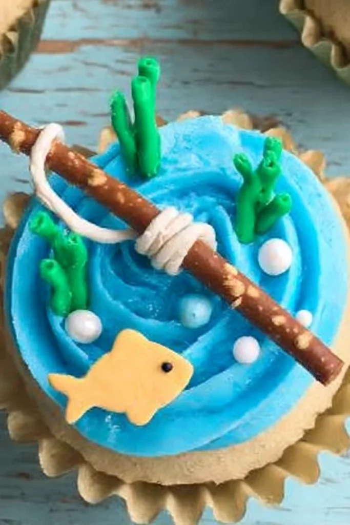 Fishing-themed cupcake with blue frosting, pretzel rod, goldfish cracker, and green fondant plants.