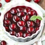 Glossy homemade cherry pie filling in a white bowl on a rustic wooden table with fresh cherries and a polka-dot napkin.