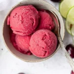 Three scoops of fresh cherry sorbet in a ceramic bowl with spoon and lime garnish