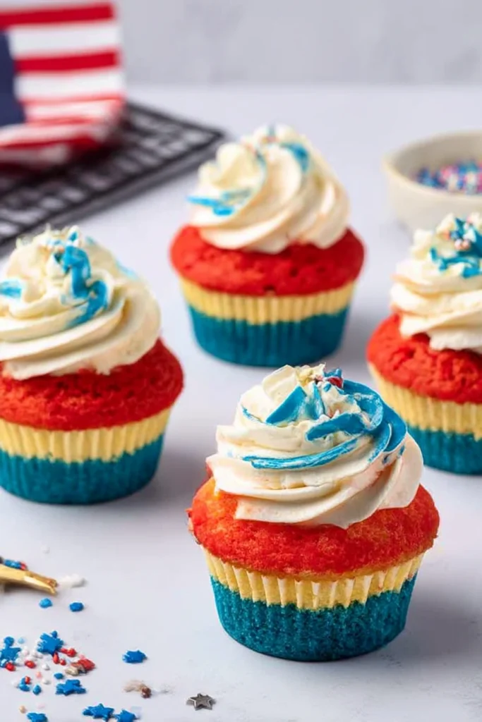 4th of July Cupcakes: Festive, Patriotic, and Simply Delicious! 5 Patriotic 4th of July cupcakes with red, yellow, and blue layers topped with swirled white and blue cream cheese frosting and festive red, white, and blue sprinkles on marble surface and cooling rack.