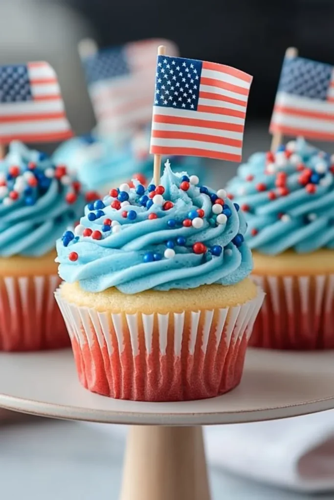 Patriotic Bomb Pop cupcakes with swirled blue frosting, festive sprinkles, and mini American flags, styled on a white stand.