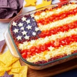 Flag Taco Dip: Easy No-Bake 4th of July Appetizer 3 Flag Taco Dip styled like the American flag with blue corn chips, cheese stars, and tomato and cheese stripes in a glass dish for a 4th of July party