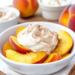 A white bowl filled with sliced ripe peaches topped with cinnamon coconut whipped cream, with whole peaches and cinnamon sticks in the background.