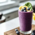 Glass of thick berry banana spinach smoothie on rustic wooden kitchen counter, topped with banana slices and blueberries.