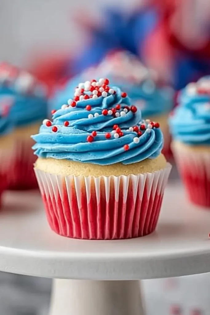 Side view of Patriotic Bomb Pop cupcakes with red-tinted bases, blue frosting, and red and white sprinkles on a cake stand.