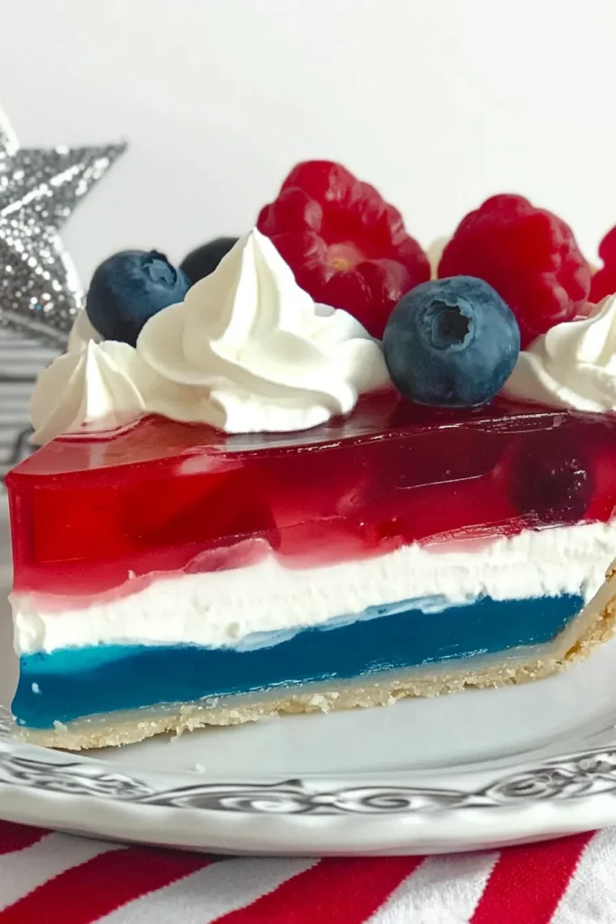 4th of July Patriotic Jello Pie: A No-Bake Red, White & Blue Treat 7 Slice of 4th of July Patriotic Jello Pie with red, white, and blue layers on a white plate, topped with whipped cream and fresh berries.