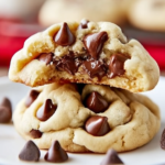 Close-up of two soft, thick homemade chocolate chip cookies stacked on a white plate, with gooey melty chocolate chips inside and scattered chocolate chips on a rustic wooden table.