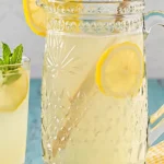 Three Ingredient Homemade Lemonade | Quick & Refreshing 5 Vintage-style glass pitcher filled with three ingredient homemade lemonade, garnished with lemon slices, next to a glass of lemonade on a rustic table.
