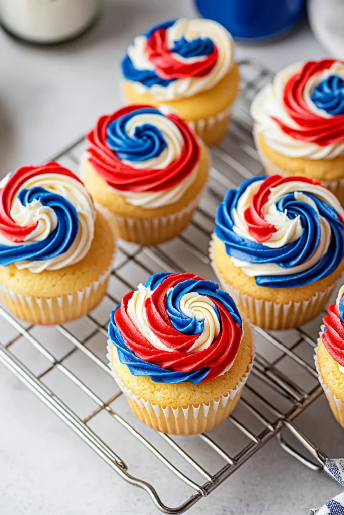 Overhead close-up of patriotic cupcakes with red, white, and blue swirled buttercream frosting on golden vanilla bases, arranged on a silver cooling rack over a gray concrete table.