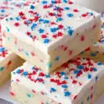 Patriotic Sugar Cookie Bars – Easy 4th of July Dessert Recipe 14 Patriotic sugar cookie bars with white frosting and red, white, and blue star sprinkles stacked on a cooling rack.
