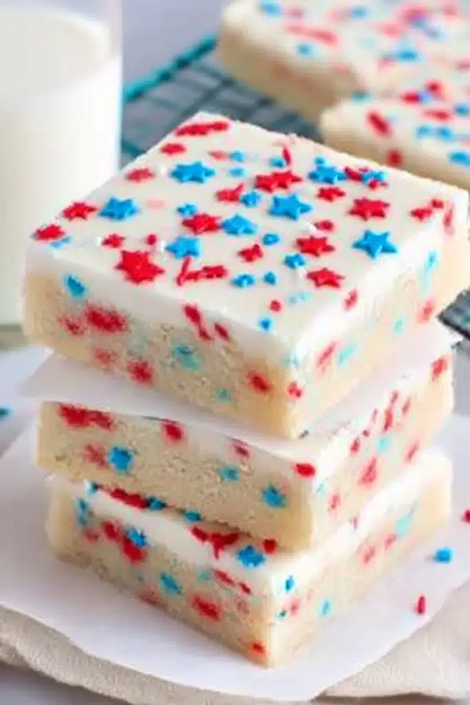 Patriotic Sugar Cookie Bars – Easy 4th of July Dessert Recipe 13 Stacked patriotic sugar cookie bars with white frosting and colorful sprinkles, on parchment paper with milk in the background.