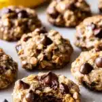 Banana Oatmeal Cookies – Soft, Healthy & Totally Delicious 12 Soft banana oatmeal cookies with chocolate chips on a light surface, one cookie bitten to show gooey chocolate center.