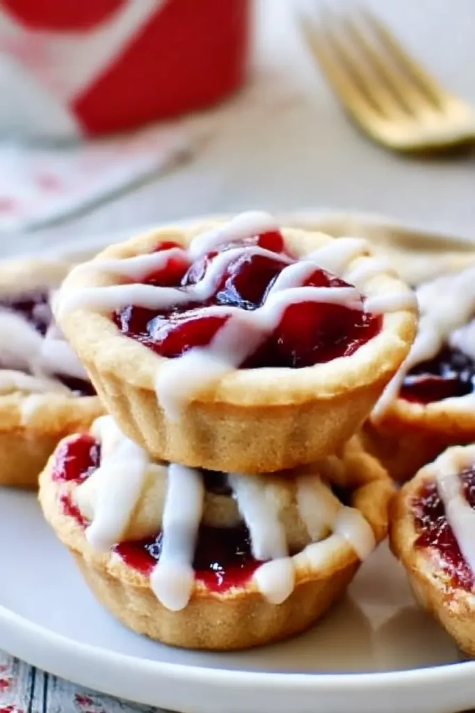 Cherry Pie Cookie Cups – Easy Bite-Sized Party Treats 6 Cherry pie cookie cups stacked on a white plate with red cherry filling and white icing drizzle