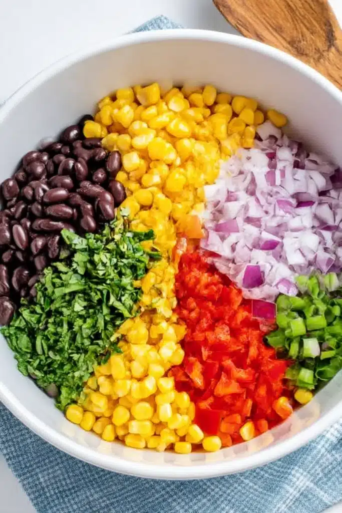 Chopped black beans, corn, bell peppers, onions, scallions, and cilantro arranged in a white bowl on a blue-check cloth

