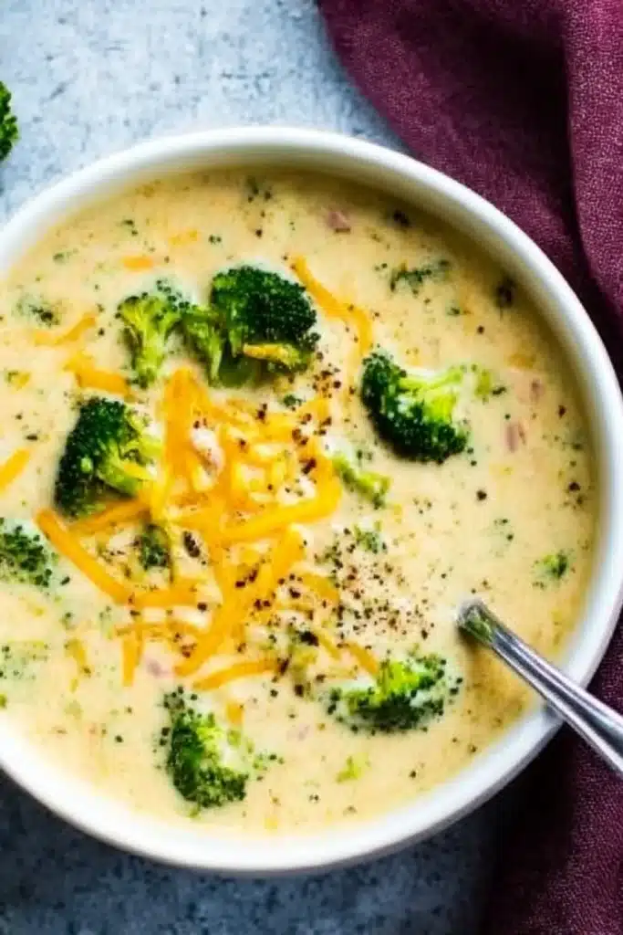 Overhead view of a bowl of creamy broccoli cheddar soup with broccoli florets, cheddar shreds, and a spoon, served on a gray background.