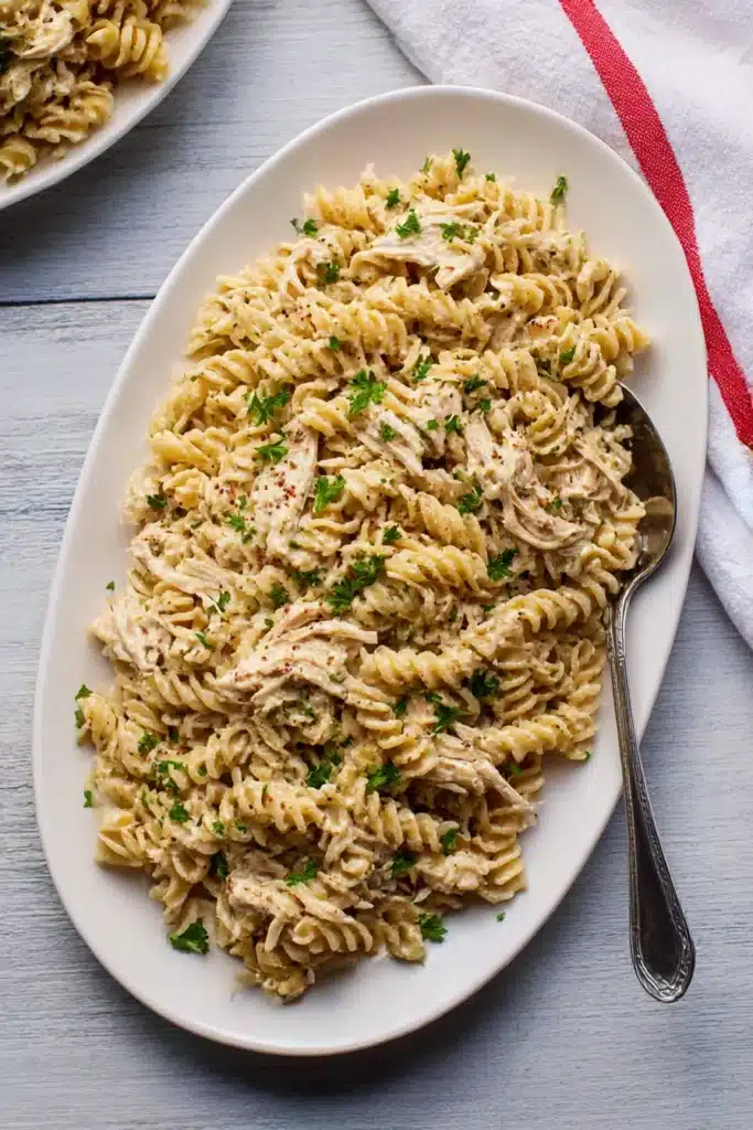 Large serving platter of creamy garlic parmesan chicken pasta with parsley, on rustic wood background with a silver spoon and kitchen towel

