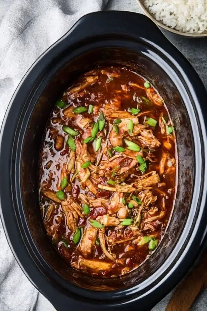 Crock Pot Bourbon Chicken simmering in a slow cooker, shredded and coated in sticky sweet sauce, garnished with green onions.