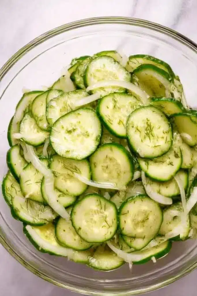 Glass bowl of cucumber vinegar salad with sliced onions and dill on a light background.

