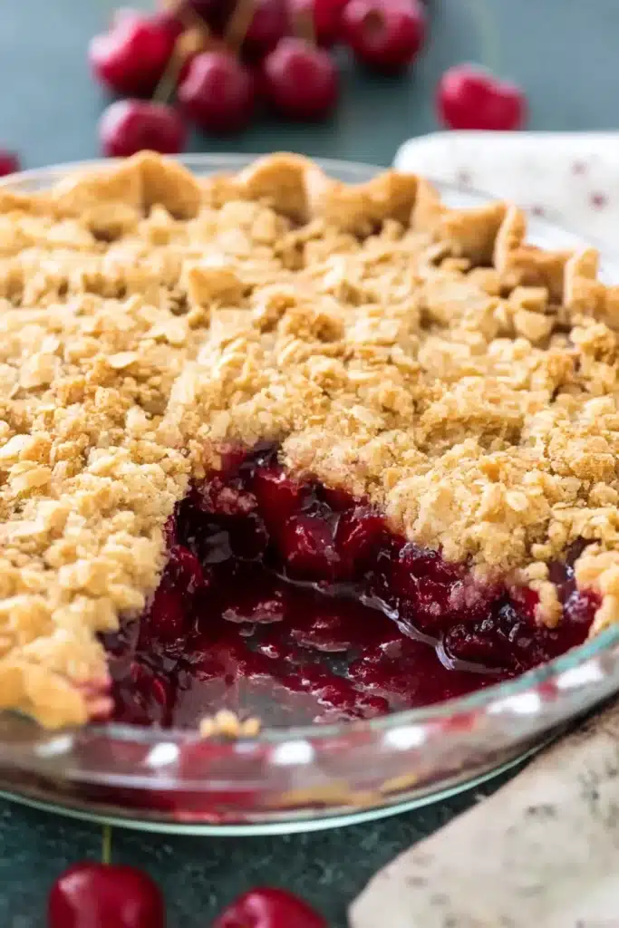 Cherry crumble pie in a glass dish with a slice missing, revealing juicy cherry filling and golden oat topping

