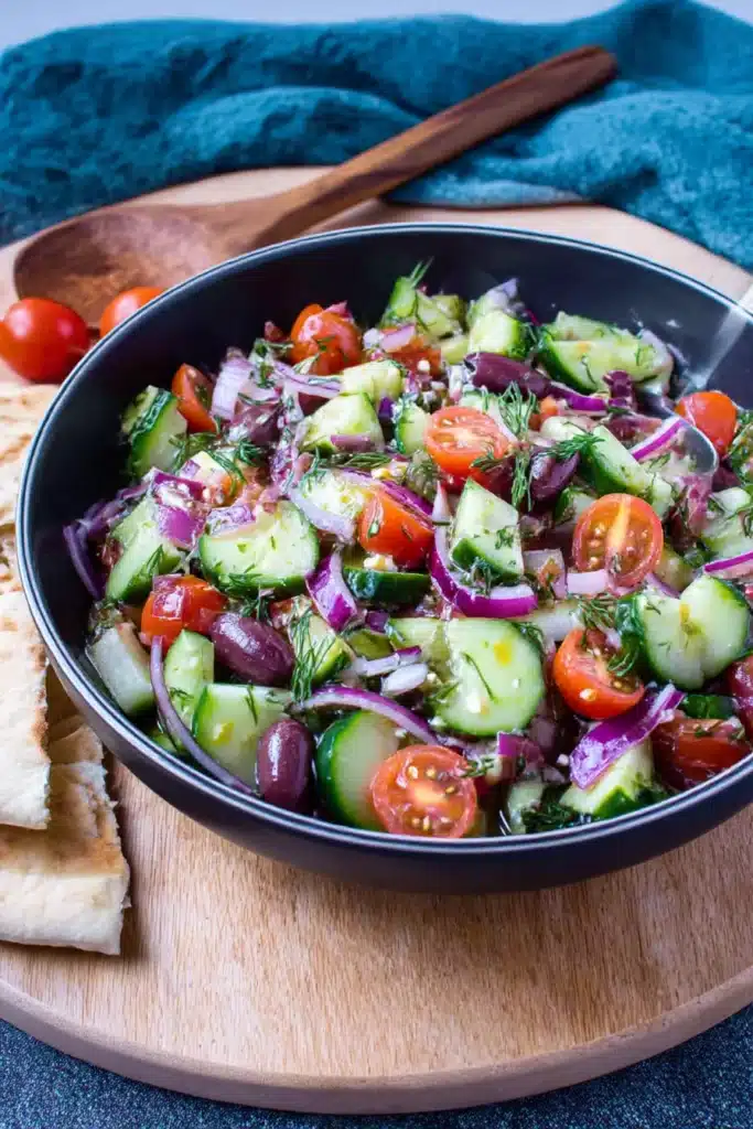 Greek cucumber salad with cherry tomatoes, red onions, and olives in a black bowl with pita on the side


