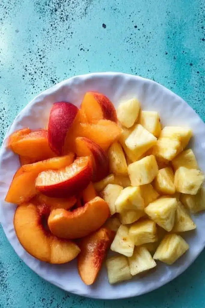 White plate with fresh sliced peaches and pineapple chunks on a teal rustic background

