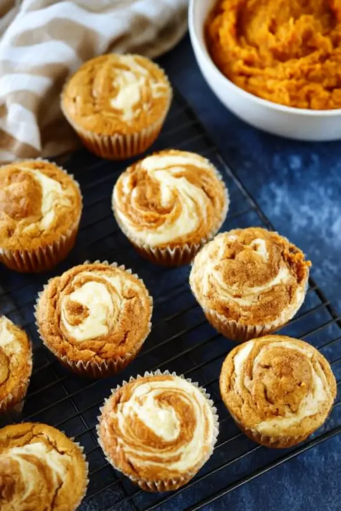 Pumpkin cream cheese muffins with creamy swirled tops on a cooling rack beside a bowl of pumpkin purée.