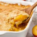 Peach Cobbler with Cake Mix – Easy 4-Ingredient Summer Dessert 7 Freshly baked peach cobbler with cake mix being scooped with a wooden spoon from a white ceramic dish, featuring golden crisp topping and juicy peaches