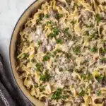 Creamy ground beef stroganoff with egg noodles in a beige skillet, garnished with parsley on a stone background.