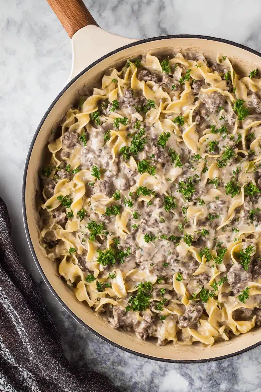 Creamy ground beef stroganoff with egg noodles in a beige skillet, garnished with parsley on a stone background.