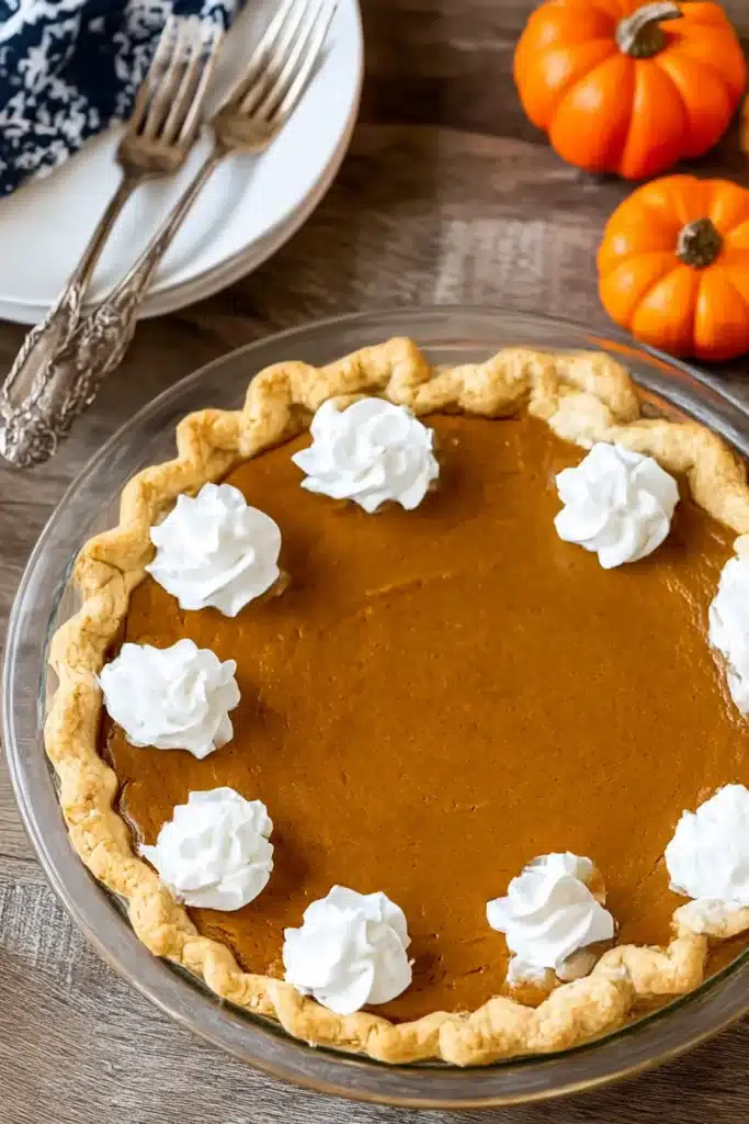 Easy Pumpkin Pie Recipe – Absolutely Delicious & Foolproof 8 Whole pumpkin pie topped with whipped cream swirls in a glass dish on a rustic table