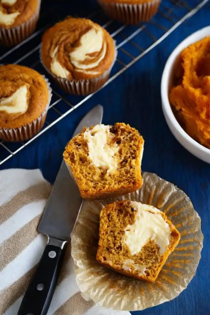 Halved pumpkin cream cheese muffin showing moist crumb and creamy swirl, on dark wood surface with knife and pumpkin purée.

