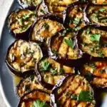 Grilled Eggplant with Garlic & Herbs (Mouthwatering & Easy) 6 Grilled eggplant slices with garlic and herbs on a white platter, topped with fresh parsley and char marks